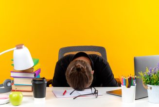 young guy student sitting at table with school tools lowered head on table isolated on orange background